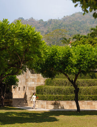 Stone sculpture beneath mature trees at Amanjiwo, with forested hillside beyond.