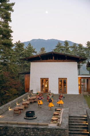 Bhutanese monks in saffron robes gather in the courtyard of Amankora's Thimphu lodge during a cultural programme, with mountains rising beyond.