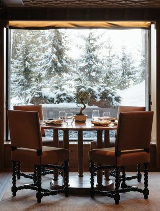 Dining table and chairs positioned by a window overlooking snow-covered pine trees at Aman Le Mélézin resort.