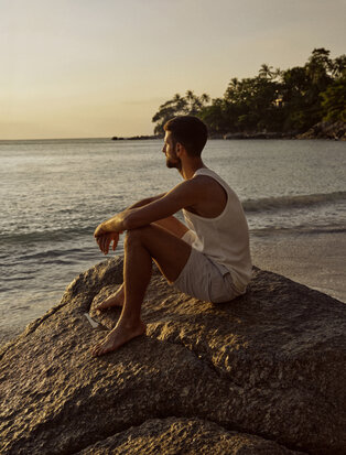 Novak Djokovic sits on a rock at Amanpuri's beach at sunset, gazing towards the sea.