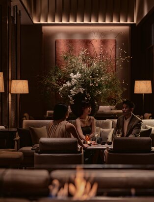 Warm-lit lounge at Aman Nai Lert Bangkok with seated guests facing a floral artwork, flanked by table lamps and fireplace glow.