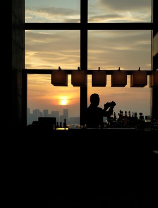 Silhouetted figures at a bar counter at Aman Tokyo, framed by a window overlooking a golden sunset over the city.