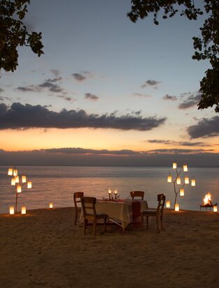 Candlelit dining setup on a beach at sunset, with two chairs facing the sea at Amanwana, Indonesia.
