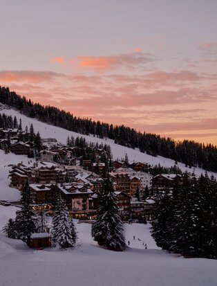 Aman Le Mélézin resort exterior at dusk, snow-covered alpine landscape beneath pink-tinted sky.