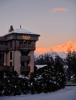 Aman Le Mélézin resort exterior at dusk, snow-covered grounds and mountains glowing in winter light.