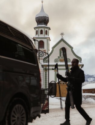 Skier boarding a shuttle at Aman Rosa Alpina, with alpine chapel visible behind in snowy Italian mountain setting.