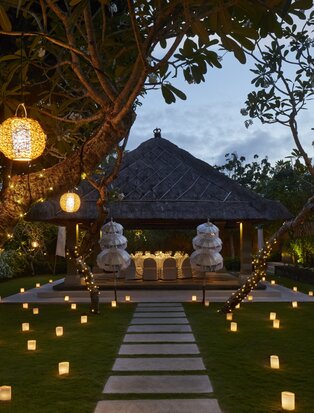 Aman Villas at Nusa Dua villa courtyard at dusk, lanterns illuminating a pathway lined with glowing candles towards a thatched-roof structure.
