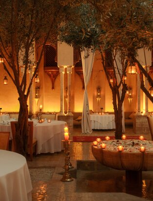 Moroccan restaurant at Amanjena lit by candlelight and warm lanterns among argan trees at dusk.