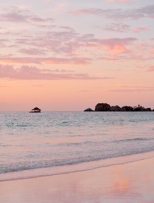Sunset view from the beach at Amanpulo, Philippines, with soft pink and orange light reflecting on the sea and sand.