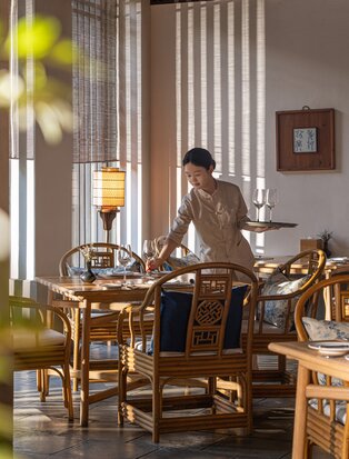Resident at Amanfayun seated at wooden desk beside tall windows with vertical blinds, warm natural light streaming through.