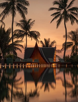 Amanpuri villa reflected in still water at sunset, framed by palm trees.