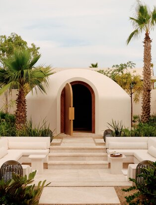 Amanvari temezcal spa entrance with curved white adobe archway and palm trees.
