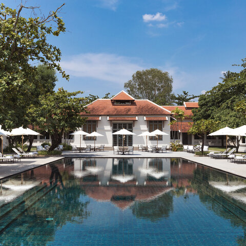 Amantaka's terracotta-coloured main pavilion reflected in a still plunge pool, framed by manicured gardens and white sun loungers under clear skies.