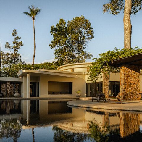 Amansara's exterior reflected in still water, with palm trees and stone architecture framed by clear sky.