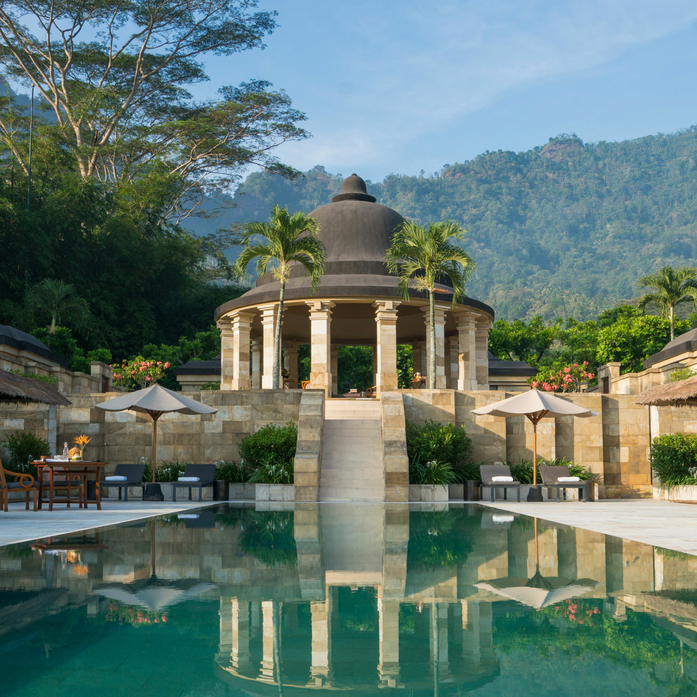 Amanjiwo's stone pavilion reflected in turquoise pool, forested mountains beyond.