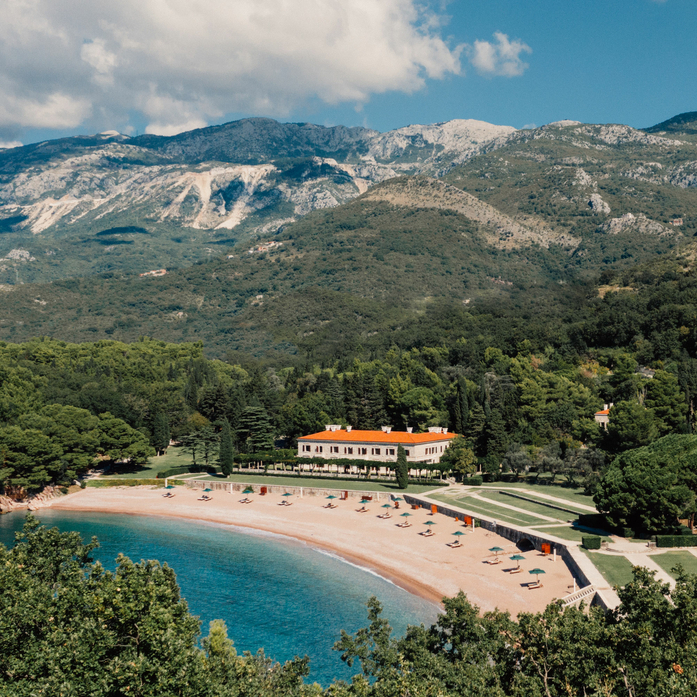 Aerial view of Aman Sveti Stefan's terracotta-roofed resort set on a peninsula, surrounded by pine forests and a curved sandy beach along the Adriatic coast.