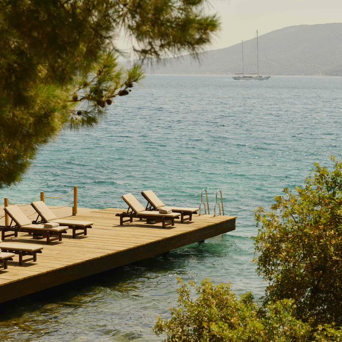 Wooden jetty with sunloungers extending into turquoise water at Amanruya, framed by pine trees.