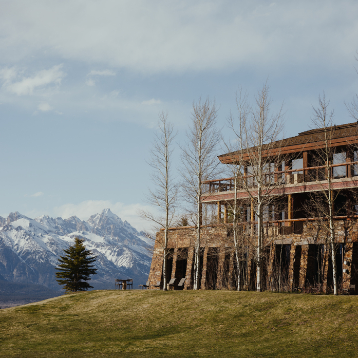 Amangani residence set on a hillside with snow-capped mountains in the distance, tall bare trees and alpine meadow in foreground.