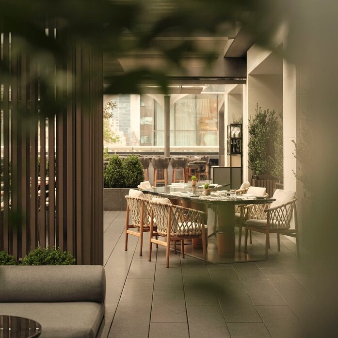 Terrazza del giardino presso Aman New York, con tavoli in legno e sedie circondate da verdure.
