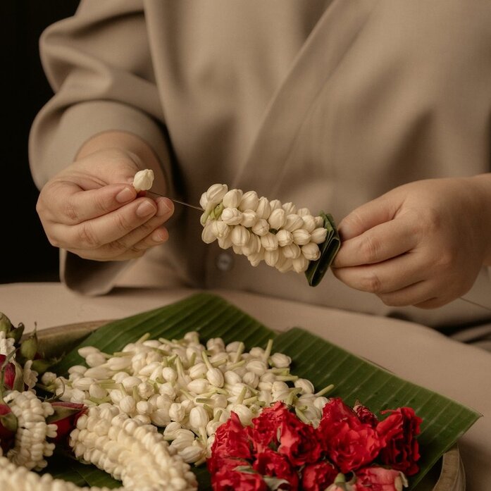 Hands holding a white flower garland at Aman Nai Lert Bangkok, with red and white blooms arranged below.