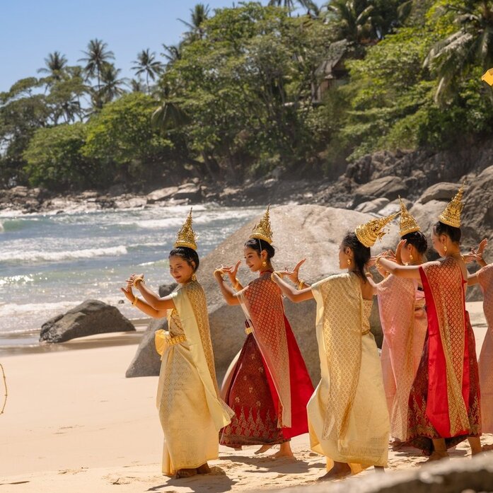 Buddhist monks in saffron and maroon robes standing on a sandy beach at Amanpuri, Thailand, with tropical palms and turquoise waters behind them.