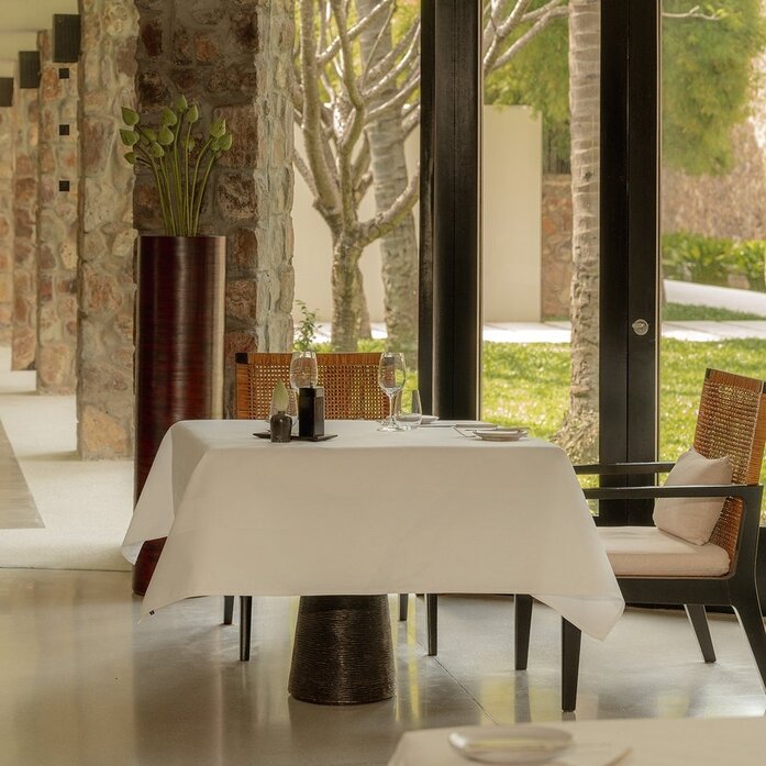 Dining room at Amansara with round table, soft furnishings and views to the gardens beyond.