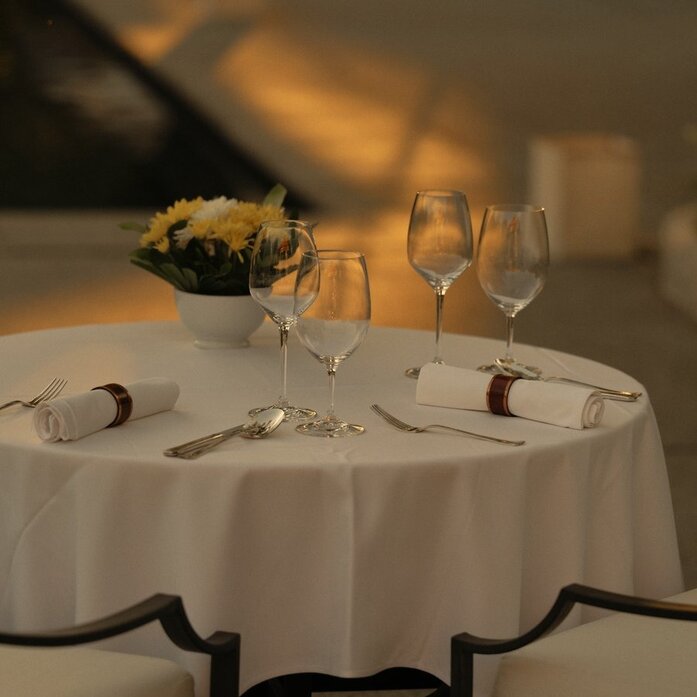 Poolside dining table at Amantaka set for an intimate dinner, with white tablecloth, place settings, and golden flowers.
