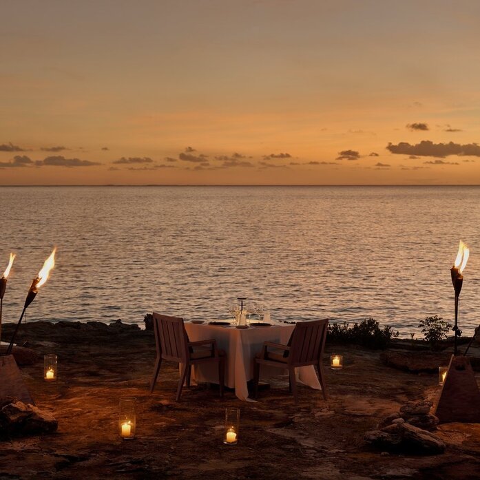 Private dining setup on the beach at Amanyara at sunset, with candlelit table overlooking calm waters.
