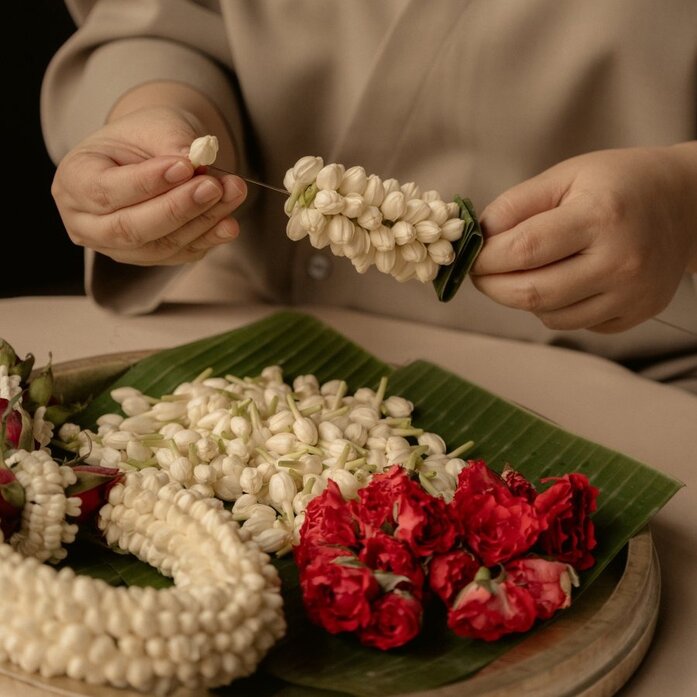 Hands holding a jasmine flower garland at Aman Nai Lert Bangkok, with finished garlands displayed on a green leaf.