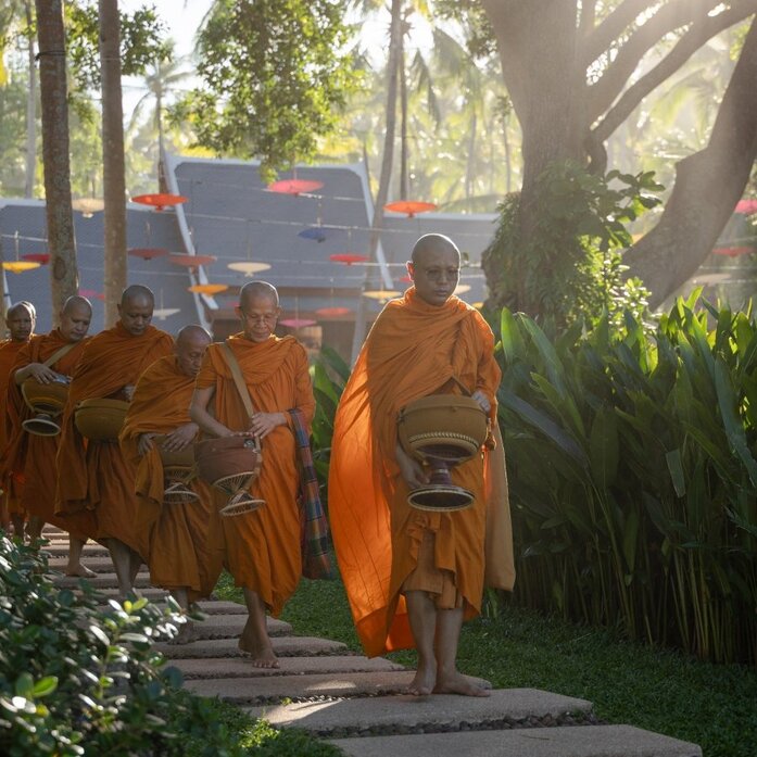 Buddhist monks in saffron robes walking through tree-lined grounds at Amanpuri during Songkran festivities.