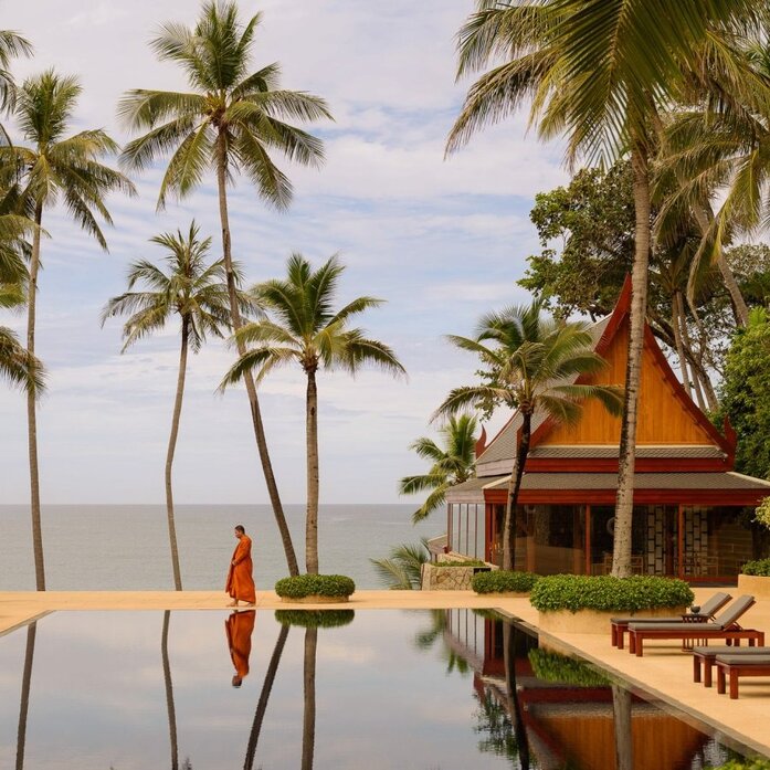 Buddhist monk in saffron robes walks along edge of infinity pool at Amanpuri, Thailand, with palm trees and golden temple architecture visible.