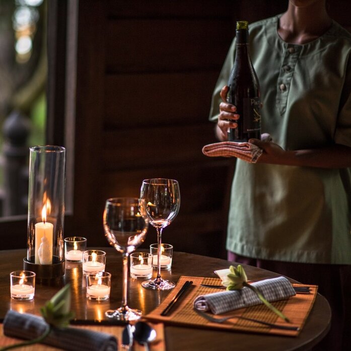 Twilight dinner setup at Amansara with candlelit table and server pouring wine in traditional Khmer village setting.