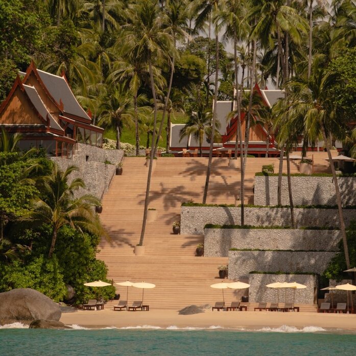 Amanpuri's beachfront exterior with wooden stairs descending to sand, flanked by tropical palms and traditional Thai architecture.