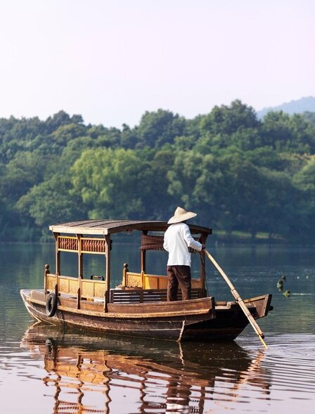 木质传统船只在西湖水面上，远处群山环绕，法云安缦度假村景色。