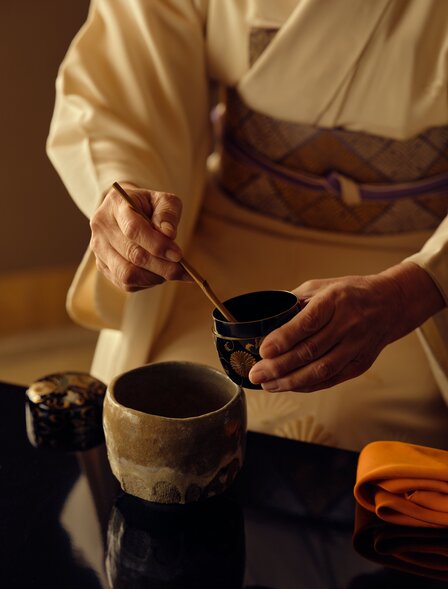 Person in traditional Japanese clothing preparing tea at Aman Kyoto tea house, holding ceramic bowl and whisk.