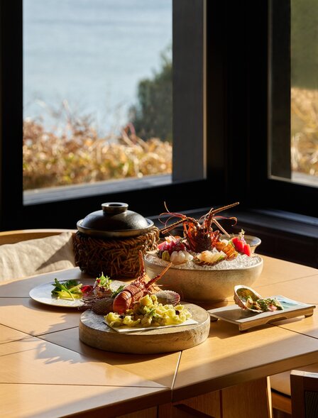 安缦伊沐度假村餐厅内，木质餐桌上摆放着精心烹饪的餐点，大窗户框景日本秋季景观。