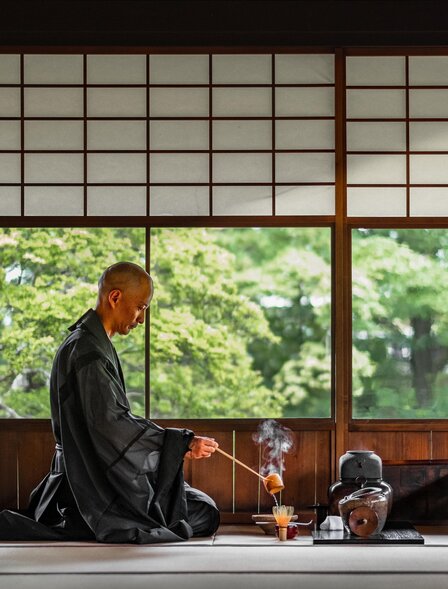 Person in dark kimono practising tea ceremony at Aman Kyoto, with garden visible through shoji screens.