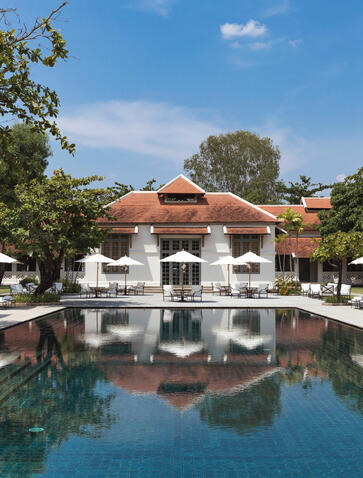 Portrait of Amantaka's principal residence reflected in still water, surrounded by lush gardens beneath clear skies.