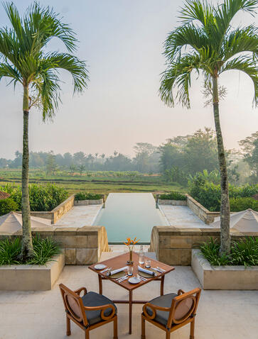 Two wooden chairs facing a plunge pool at Amanjiwo, flanked by palm trees overlooking distant temples.