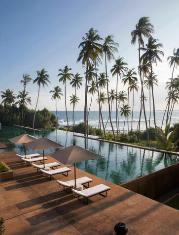 Amanjiwo's infinity pool overlooking palm-lined waterways, with wooden loungers and clear blue sky.