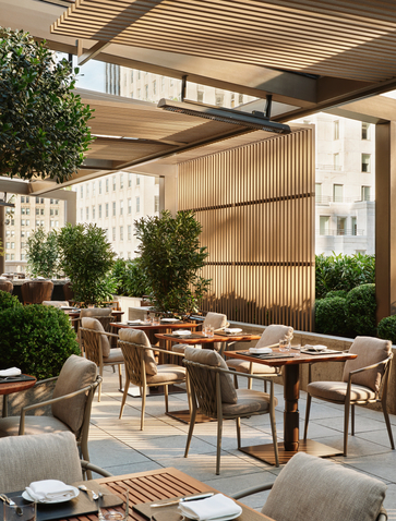 Garden terrace at Aman New York with wooden dining table, chairs, and potted plants under a slatted canopy.
