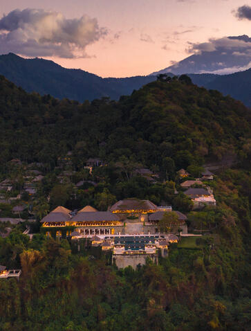 Amankila resort nestled amongst forested hills at dusk, with warm-lit buildings set against mountain peaks under a soft purple sky.