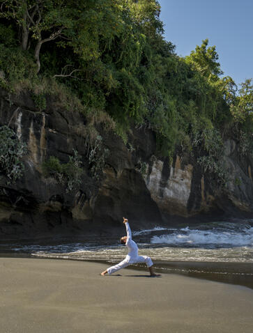 Person practising yoga on a sandy beach at Amankila, Indonesia, with forested cliffs rising behind.