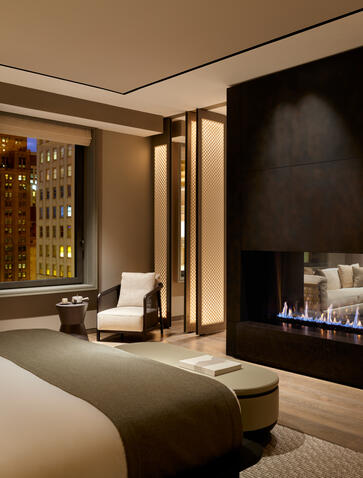 Aman Suite at Aman New York with bed, fireplace, and contemporary furnishings in warm neutral tones.