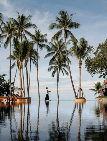 Staff member serving drinks by the swimming pool at Amanpuri, Thailand, with palm trees reflected in still water.
