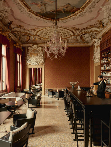 Bar at Aman Venice with ornate ceiling, warm terracotta walls, and period furnishings.