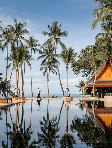 Amanpuri's infinity pool reflecting palm trees and golden-roofed pavilion under clear sky.