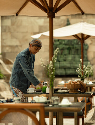 Terrace breakfast setup at Amanjiwo, with a staff member arranging flowers on a wooden table beneath umbrellas.
