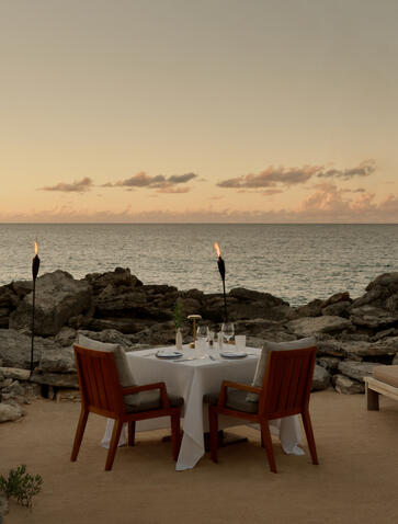 Candlelit dining table set on the beach at Amanyara at sunset, with burgundy chairs overlooking calm waters.