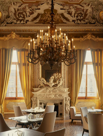 Ornate palazzo dining room at Aman Venice with gilded ceiling, crystal chandelier, and tall draped windows.